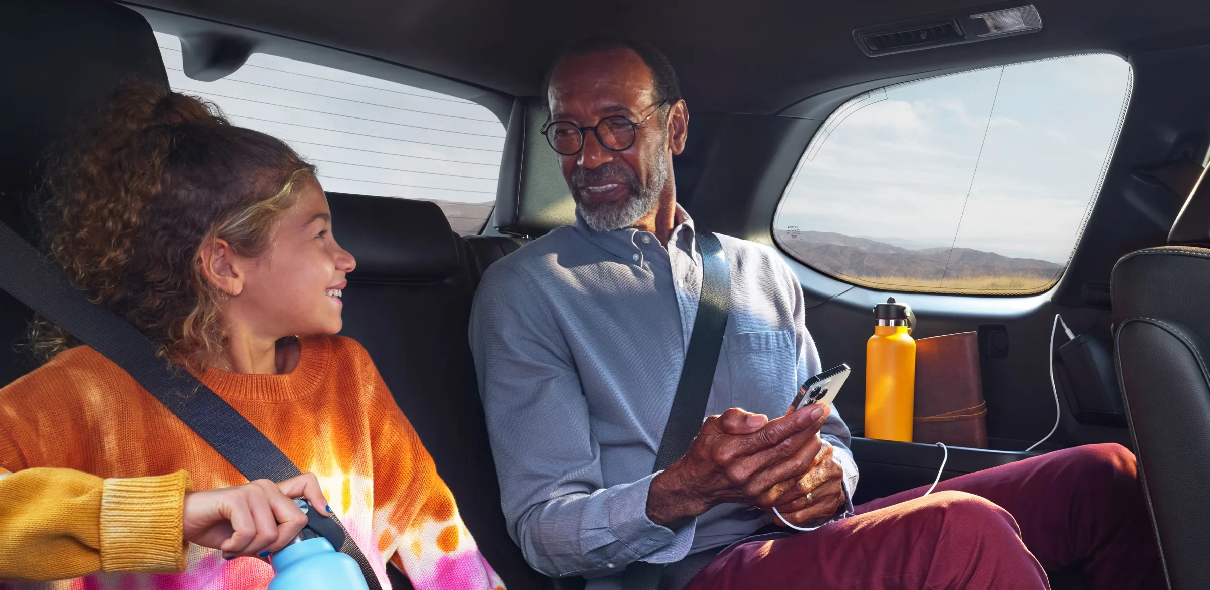 A grandfather and a smiling child side by side in the second row of a Toyota Grand Highlander, a water bottle in the door and hills outside the window.
