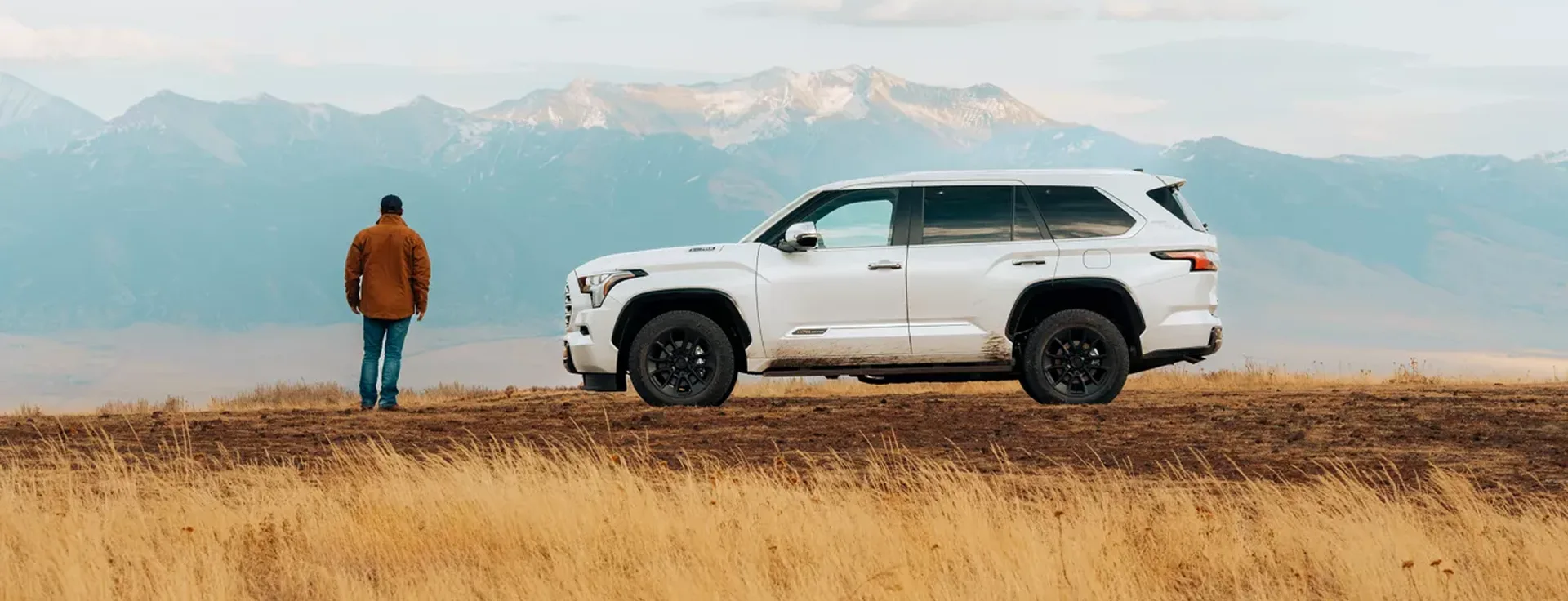 A white Toyota Sequoia parked on a grassy hillside, a man standing beside it looking out at the mountains
