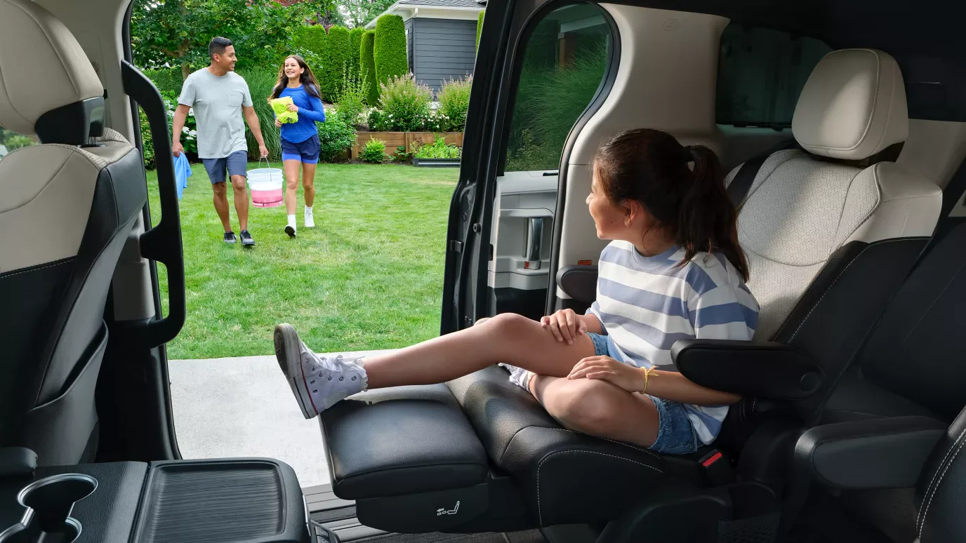 A girl in the second row of a Toyota Sienna with the sliding door open, her family playing on the lawn outside.