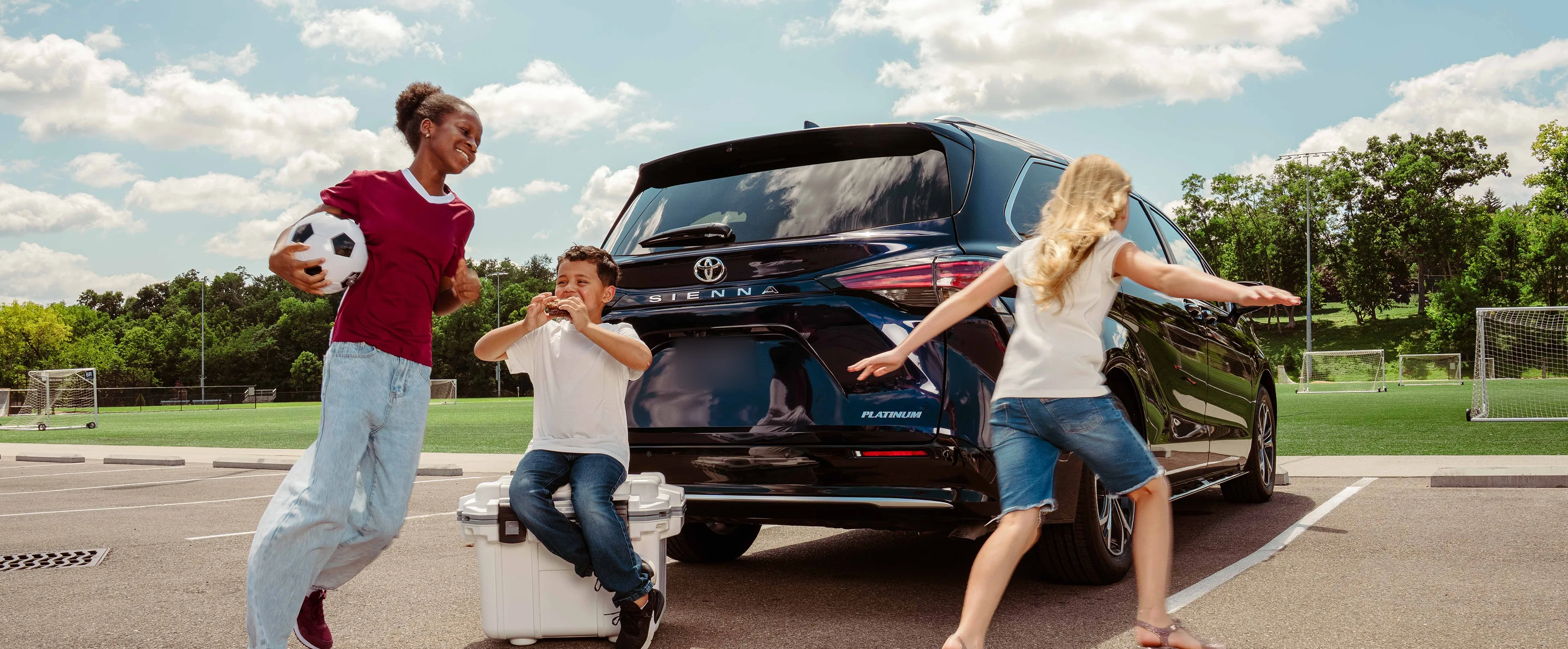 Three children playing beside a black Toyota Sienna Platinum at a soccer field, one sitting on a white cooler